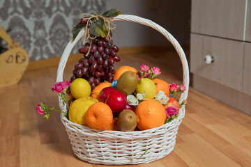 A bunch of grapes hangs from the handle of the basket. The white basket contains grapes, kiwi, orange, red apple, quince, orange, and lemon. There are pink and white golden flowers.