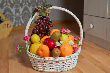 A bunch of grapes hangs from the handle of the basket. The white basket contains grapes, kiwi,...