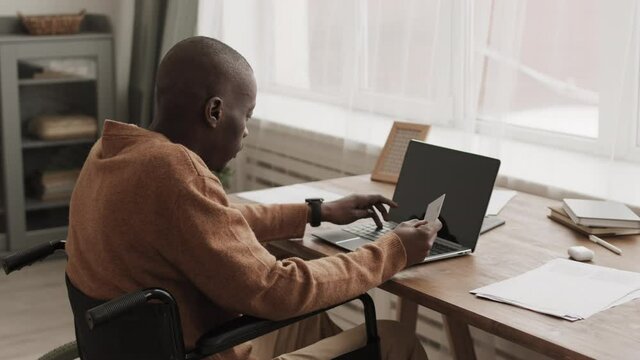 Back view of young African disabled man sitting on wheelchair at desk in living room with card in one of his hands and making online purchase