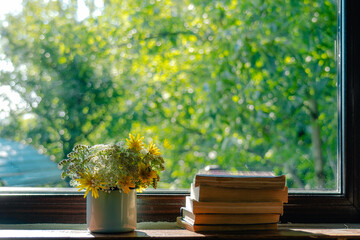 Beautiful small bouquet of simple meadow flowers in blue metal mug standing on window sill near pile of old paper books stacked together. Sunny green bokeh of leaves of trees behind glass