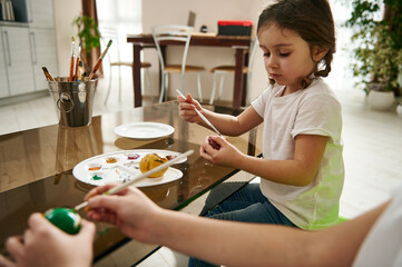 Adorable little girl with braids decorating an Easter egg, sits at the table next to her brother at home