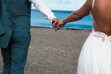 Hände von einem Braut Paar halten sich fest. Sie gehen über dem Strand an der Ostsee. Man sieht Strand und Wasser. Die Braut  hat weßes Kleid an, der Bräutigam die blaue Weste und Hose, das weiße Hemd