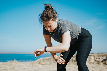 Tired young athletic woman runner looking at fitness tracker checking pulse or sport information, taking a rest at mountain landscape nature path near blue sea. Weight loss cardio goal achievement