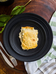 Delicious lasagna four cheeses on black ceramic plates, green leaves, cutlery and dishcloth with wooden base.
Close-up, top view.