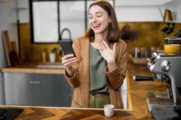 Pretty positive woman dressed in a brown jacket speaking on phone before the work in the morning, drinking coffee on stylish kitchen design.