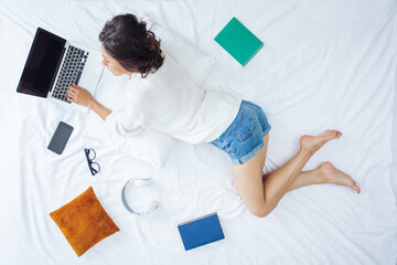 Above view full-length portrait of pretty young woman using laptop computer in her bed, working distantly from home on self-isolation