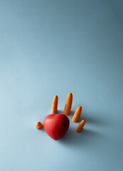 A human hand made of fresh carrots comes out of the blue background and reaches for the red heart. Health care and self care concept.  Creative minimal concept.