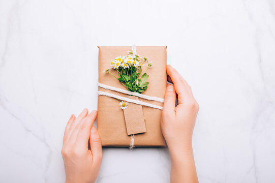 Hands are holding gift wrapped in recycled brown paper decorated with real fresh chamomiles, minimalist style wrapping, top view flat lay, eco friendly and zero waste packaging