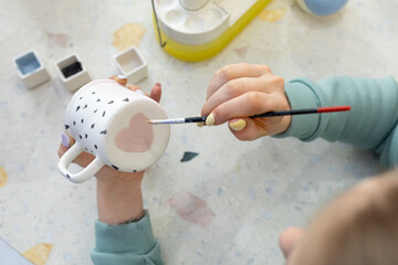 Women's hands paint pink heart on back of ceramic cup with brush. Training in profession of...