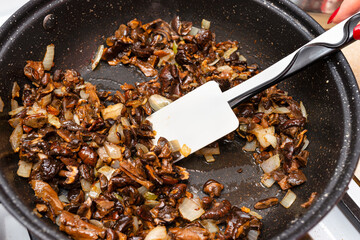A woman is holding a silicone spoon and mixing fried mushrooms with onions in a frying pan on a gas stove. © Michal