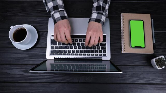 Top view of the hands of a girl who type, writes emails on an laptop. Near the laptop is a smartphone with a green screen and a chrome key. Place for your advertising content. Slow motion. Close up.