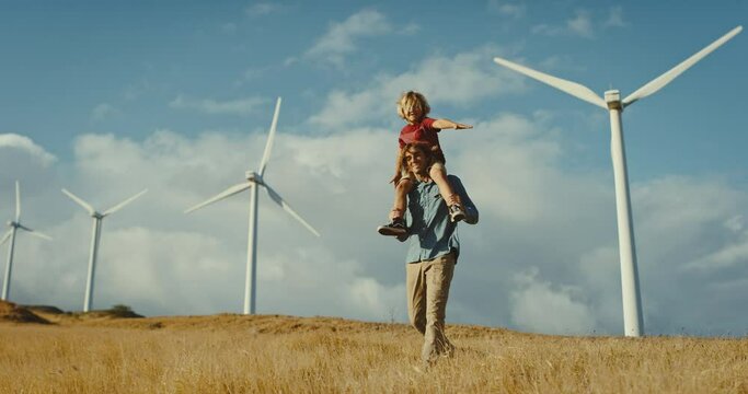 Father with young son on his back looking out at windmills on the hillside at sunset, cleaner and sustainable future, climate change, father and son bonding
