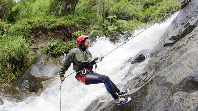 A Young Woman Slowly Rappels Down A Cliff Face Beside A Strong Flowing Cascade On Sunny Day