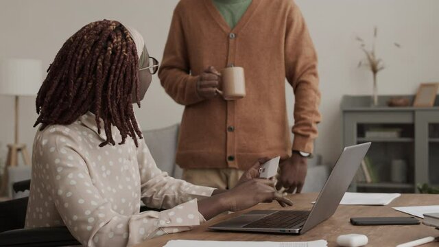 Lockdown of young African disabled woman sitting on wheelchair at laptop at desktop with card in hands and shopping online while unrecognizable man standing nearby with tea cup