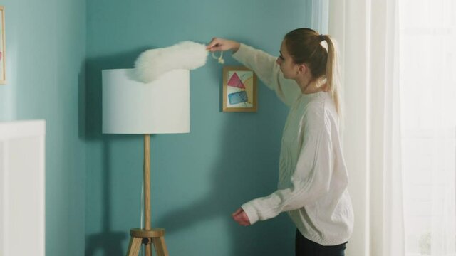 Pretty girl in a white sweater in a spacious bright room the color of a sea wave, with a window, wipes the dust from the lamp, beige sofa and white bookcase.