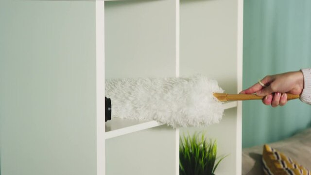 Pretty girl in a white sweater in a spacious bright room the color of a sea wave, with a window, wipes the dust from the lamp, beige sofa and white bookcase.