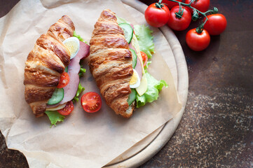 Croissant sandwiches with ham, cheese, fresh salad, tomatoes, eggs and cucumber on wooden circle board. Breakfast with fresh croissant sandwiches. Top view