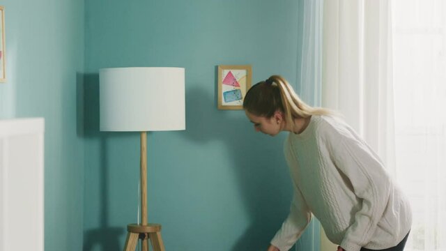 Young woman in a white sweater in a spacious bright room the color of a sea wave, with a window, wipes the dust from the lamp, beige sofa and white bookcase.