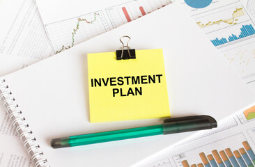 A yellow sticker with text Investment Plan is in a Notepad with a green pen financial charts and documents