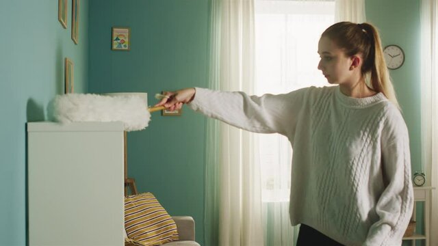Pretty girl in a white sweater in a spacious bright room the color of a sea wave, with a window, wipes the dust from the lamp, beige sofa and white bookcase.