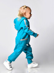 Side view of a little girl in a blue warm jumpsuit and white sneakers walking along a gray wall in the studio. The concept of advertising outerwear for children. Place for your text.