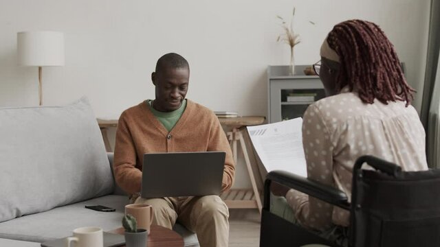 Medium shot of young African disabled woman sitting on wheelchair with document in hands and dictating text to African man sitting on sofa and typing on laptop keyboard