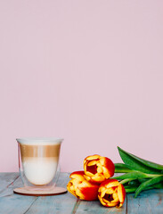 Cappuccino or latte with white foam in a double-walled glass on a round board and a bouquet of tulips in the lower right corner