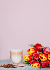 Cappuccino in a glass with double walls and roasted coffee beans with a bouquet of tulips. Feminine pink background with copy space. High resolution image, shallow depth of field.