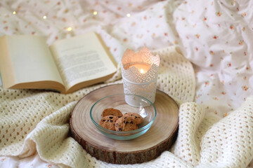 Bowl of chocolate chip cookies, lit candle and open book on a bed. Selective focus.