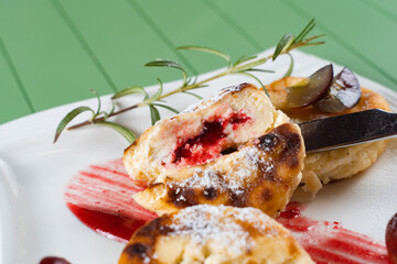 Cheesecakes cut with berries, decorated with powdered sugar, rosemary branch and grapes with berry jam on a white plate. Delicious cottage cheese breakfast.