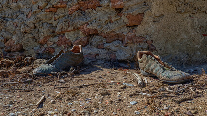 Old worn-out shoes with worn leather and no laces in an abandoned house. There is a pile of garbage around, the background is a wall of red stone and clay.