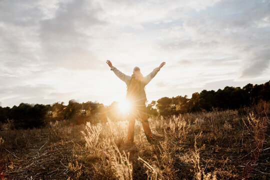 Senior male hiker with arms raised standing in filed during sunset