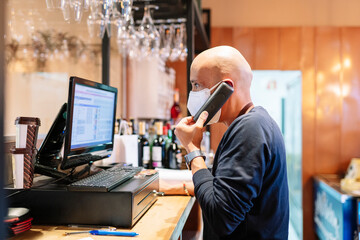 Male owner in mask taking orders through phone while standing in front of computer during pandemic