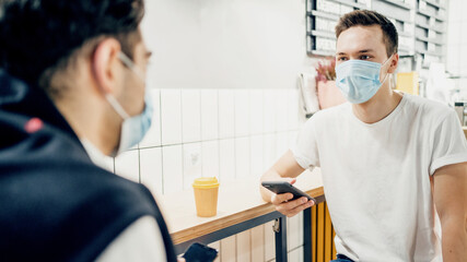 Obraz premium Men in a mask from the virus, pandemic, coronavirus, drink coffee and communicate in the messenger on the phone. The distance of 1 meter between people in a restaurant cafe. Two clients