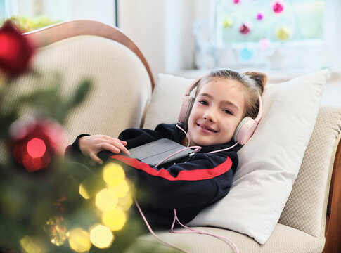 Smiling girl with digital tablet listening music through headphones while lying on sofa at home during holiday