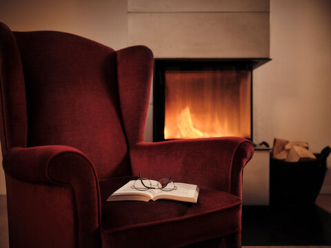 Eyeglasses with open book on maroon wingback chair by fireplace at home