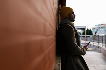 Mid adult man with mobile phone looking away while leaning on wall