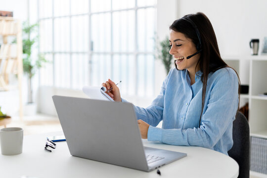 Cheerful businesswoman wearing microphone headset writing in notepad while sitting with laptop at office