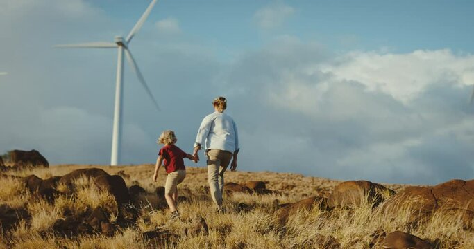 Adventurous father and son hiking up hillside with windmills, family lifestyle, quality time together, clean energy for future generations