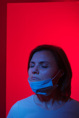 the abstract photo of a frustrated woman with closed eyes in medical mask over her chin  in a blue light over the red background