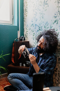 Hipster Man Holding Camera While Sitting Cross Legged Against Wall At Home