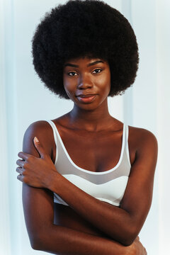 Afro fashion model in white lingerie hugging herself while standing against white wall