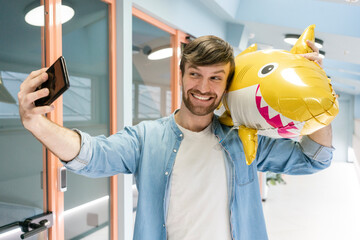 Smiling creative businessman taking selfie with balloon shark in corridor at workplace