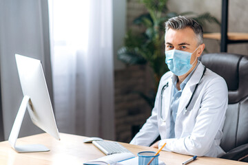 Portrait of male middle aged general doctor with gray hair dressed in lab coat and medical mask looks directly at the camera at workplace at the clinic. Doctor's consultation, health concept - Powered by Adobe