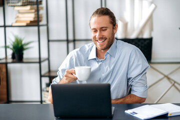 Joyful confident caucasian guy, employee or freelancer, holding a cup of coffee while sitting at his desk at home or office, looking at the camera with a friendly smile