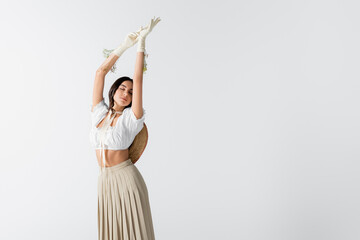 Obraz premium brunette woman in stylish spring outfit and gloves with flowers posing with hands above head isolated on white