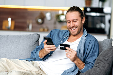 A satisfied smiling caucasian guy, in casual stylish clothes, sits on the couch at home, holds a phone and a credit card in his hands, pays for online purchases, enters a card number, online shopping