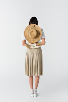 Full Length Of Woman In Gloves With Flowers And Straw Hat Behind Back On White