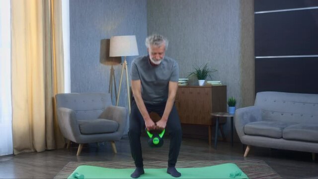 Gray-haired senior man with a beard does fitness exercise with a kettlebell. The grandfather in the living room is lifting a weight. Old man in sportswear. Success and achievement after hard work