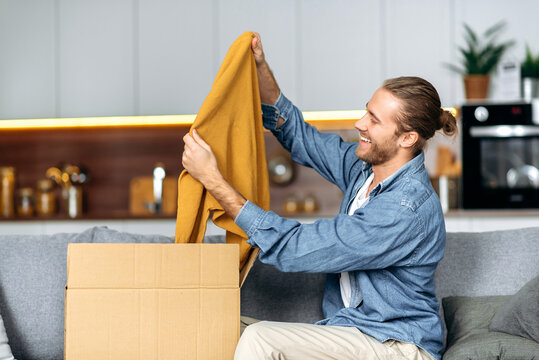 Smiling Attractive Man Unpacked His Parcel, Happy About Getting A Long Expected Order. Caucasian Modern Guy Shopping In Internet Stores, Buying New Clothes Online, Online Shopping Concept